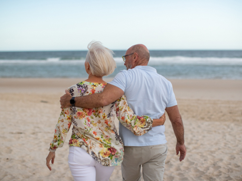 Older couple walking on beach with arms around each other | Icon Cancer Centre